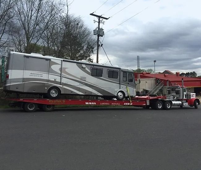 Gray RV on a red flatbed tow truck under cloudy skies by a roadside