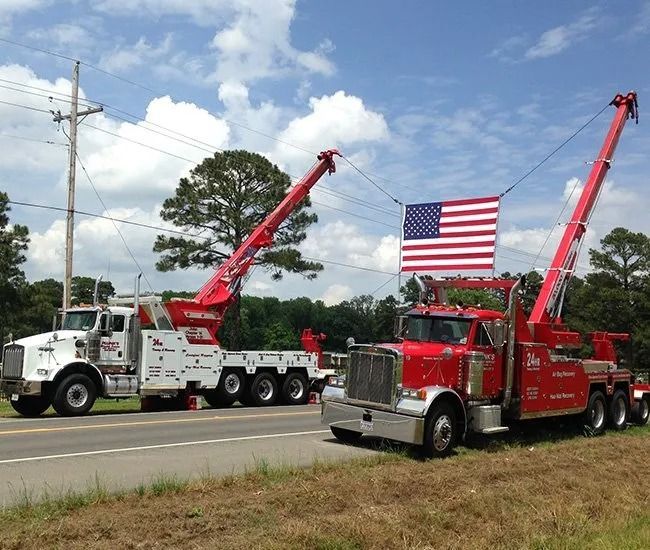 Two tow trucks with raised red booms parked roadside, one red and one white, with a large American flag behind them