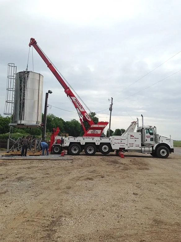 Red and white crane truck lifting at an industrial site near a silo under cloudy skies