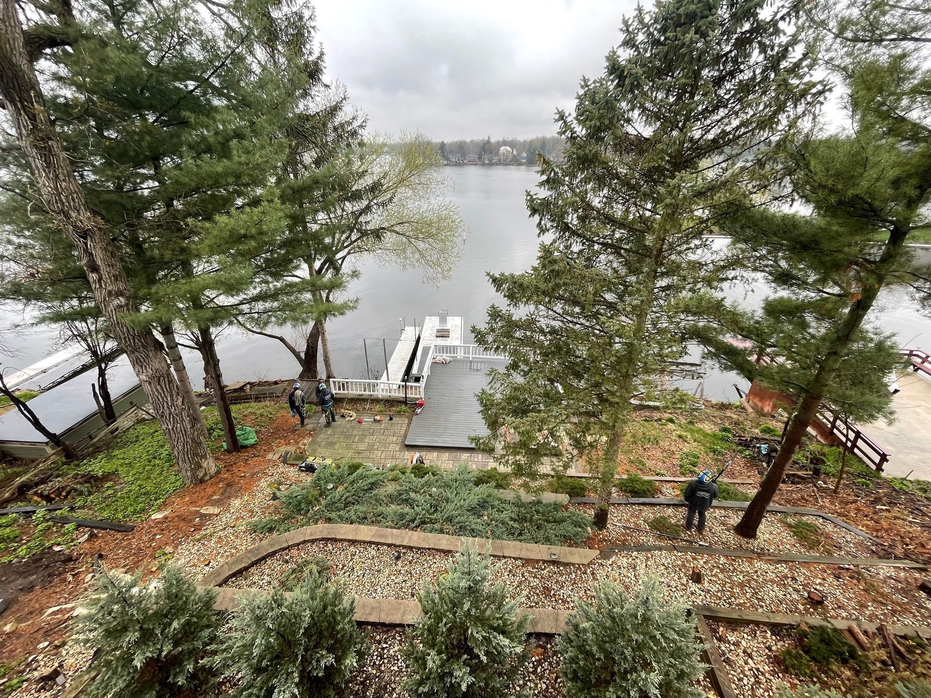 Overhead view of a lake with a dock, trees, and terraced landscaping. People are near the dock. Cloudy day.