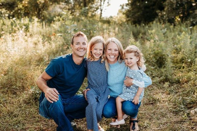Family of four, smiling and posing in field; dad in blue shirt, two daughters, mom in blue sweater.