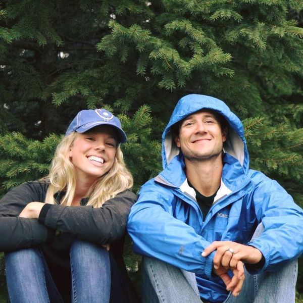 Smiling woman and man sitting outdoors, woman in cap and dark jacket, man in blue jacket hood up, evergreen trees.