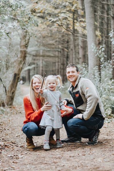 Family posing in a forest: woman in red, girl with pigtails holding a stick, man in fleece, all smiling.