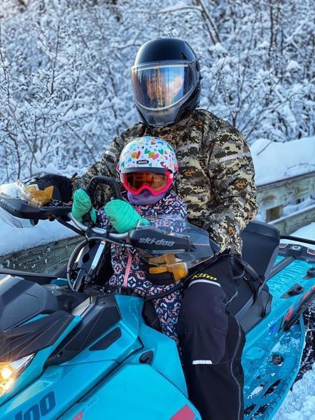 Person and child riding a teal snowmobile on a snowy day. Both wear helmets and cold-weather gear.
