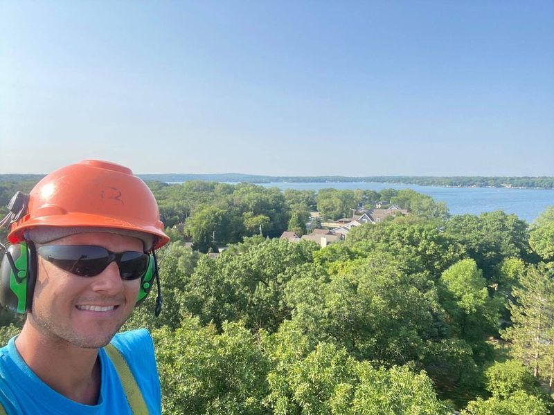 Man in orange hard hat and sunglasses, smiling, with ear protection, overlooking a tree-filled landscape and lake on a sunny day.