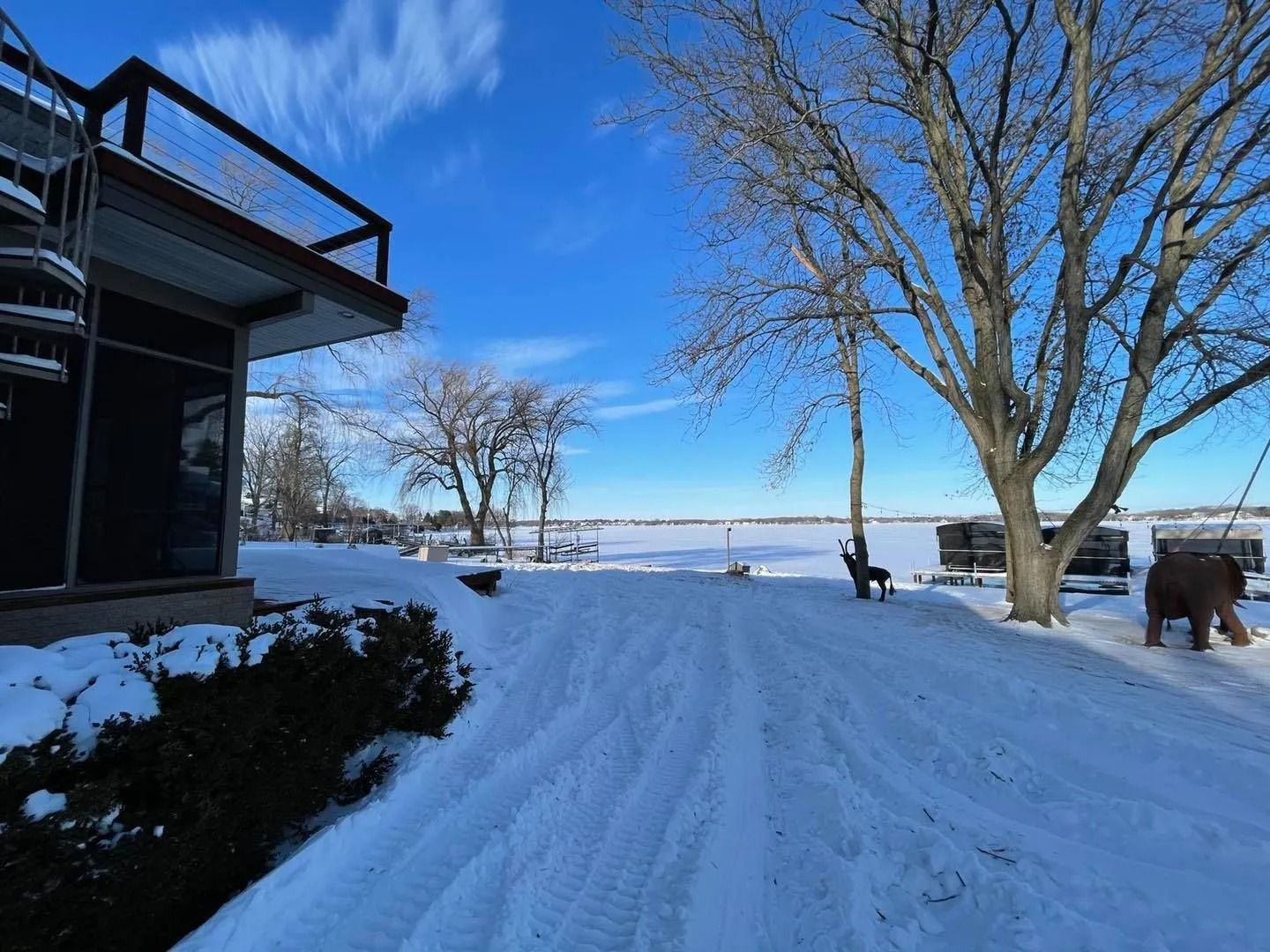Snow-covered landscape with a building on the left, trees, and a frozen lake under a bright blue sky.