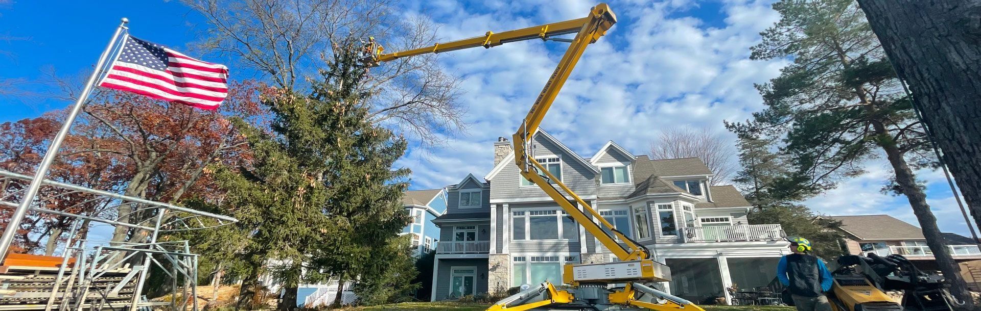 A tree trimming service is working on a tree near a multi-story house with an American flag.