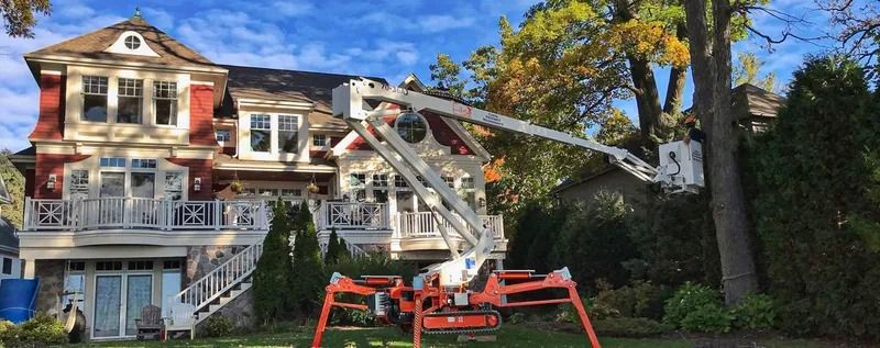 A cherry picker trims a large tree near a red and white house. Blue sky and green grass are visible.