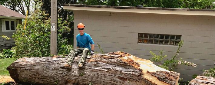 Person sitting on a large log, wearing an orange hat, sunglasses, and a blue shirt, next to a building.