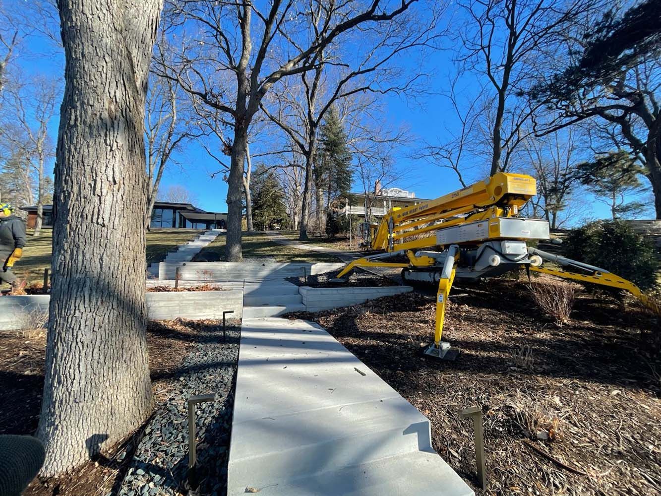 Concrete walkway construction with yellow lift machine and leafless trees.