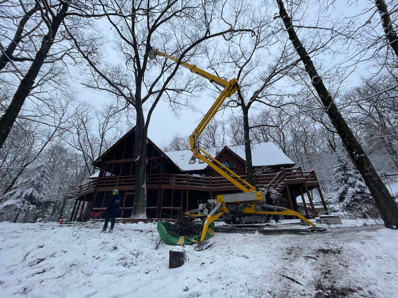 A yellow boom lift trimming a tree near a snowy cabin in a wooded area. A person stands nearby.