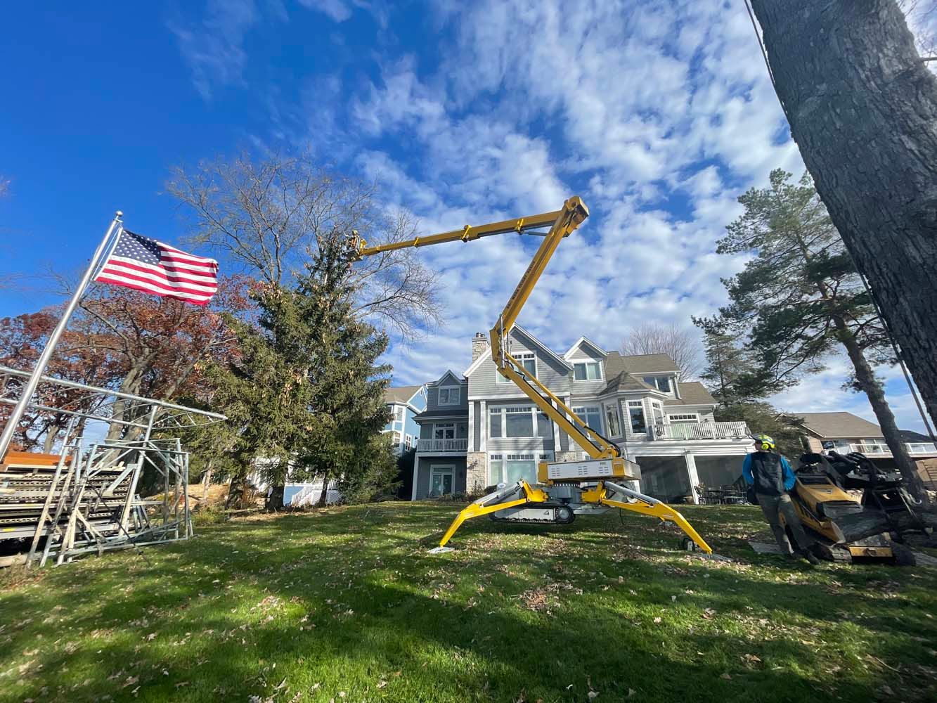 A yellow crane trimming a tree in a residential yard, American flag in background, blue sky.