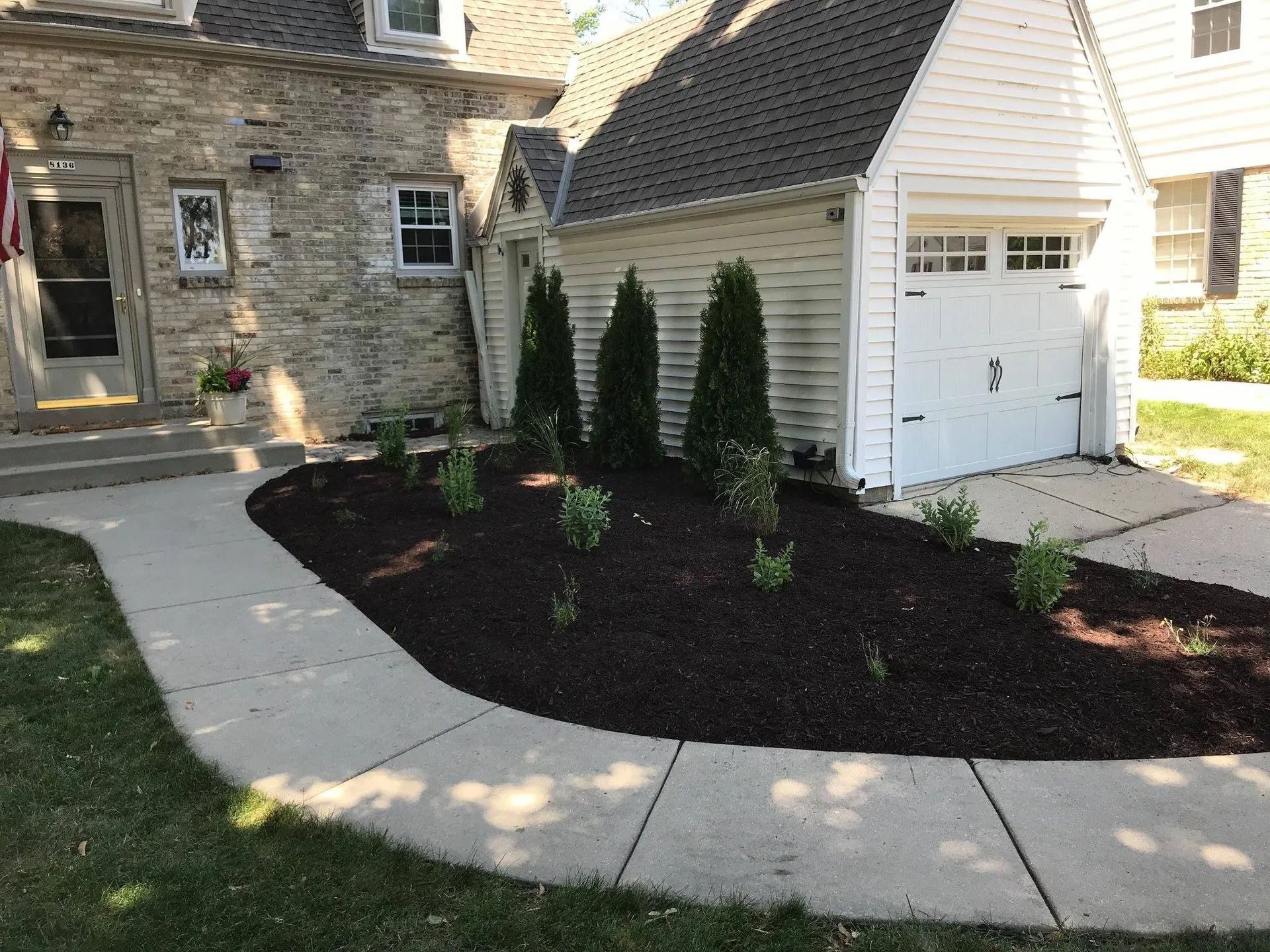 Curved walkway leading to a house with a newly mulched garden bed, containing young plants and evergreen trees.