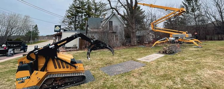 Two yellow construction machines operating near a small house. One with a claw and the other with a long arm.