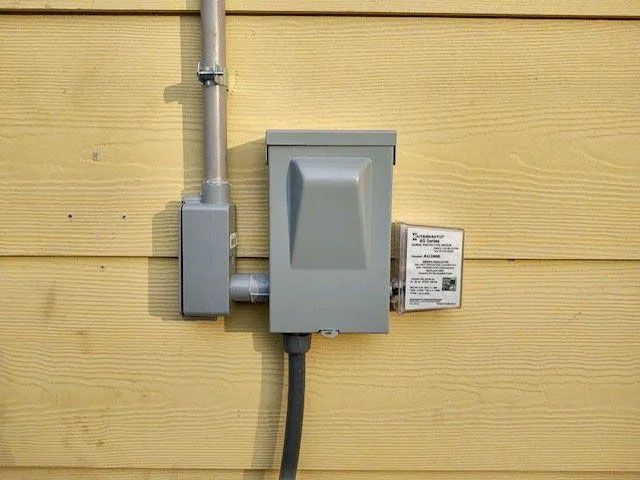 Gray electrical boxes mounted on yellow siding with metal conduit and a warning label