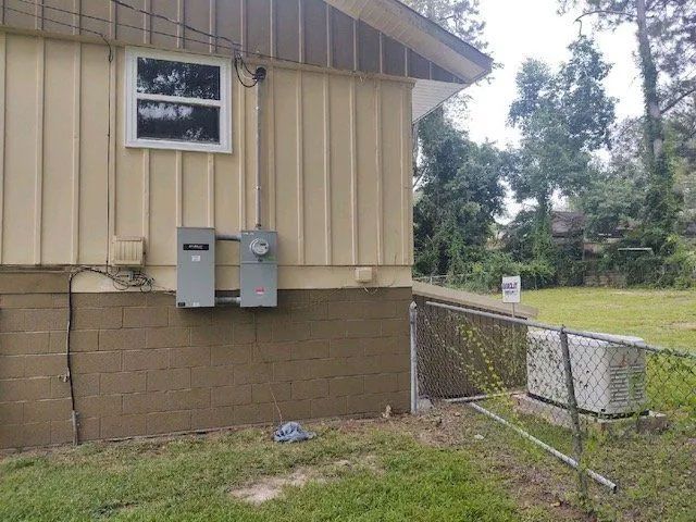 Side of a tan house with utility meters, a window, and a chain-link fence beside a grassy yard.