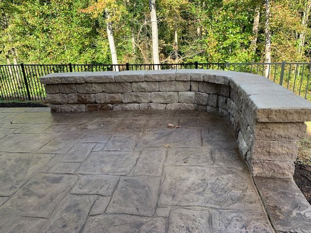 Stone patio with curved retaining wall and black railing, surrounded by trees.