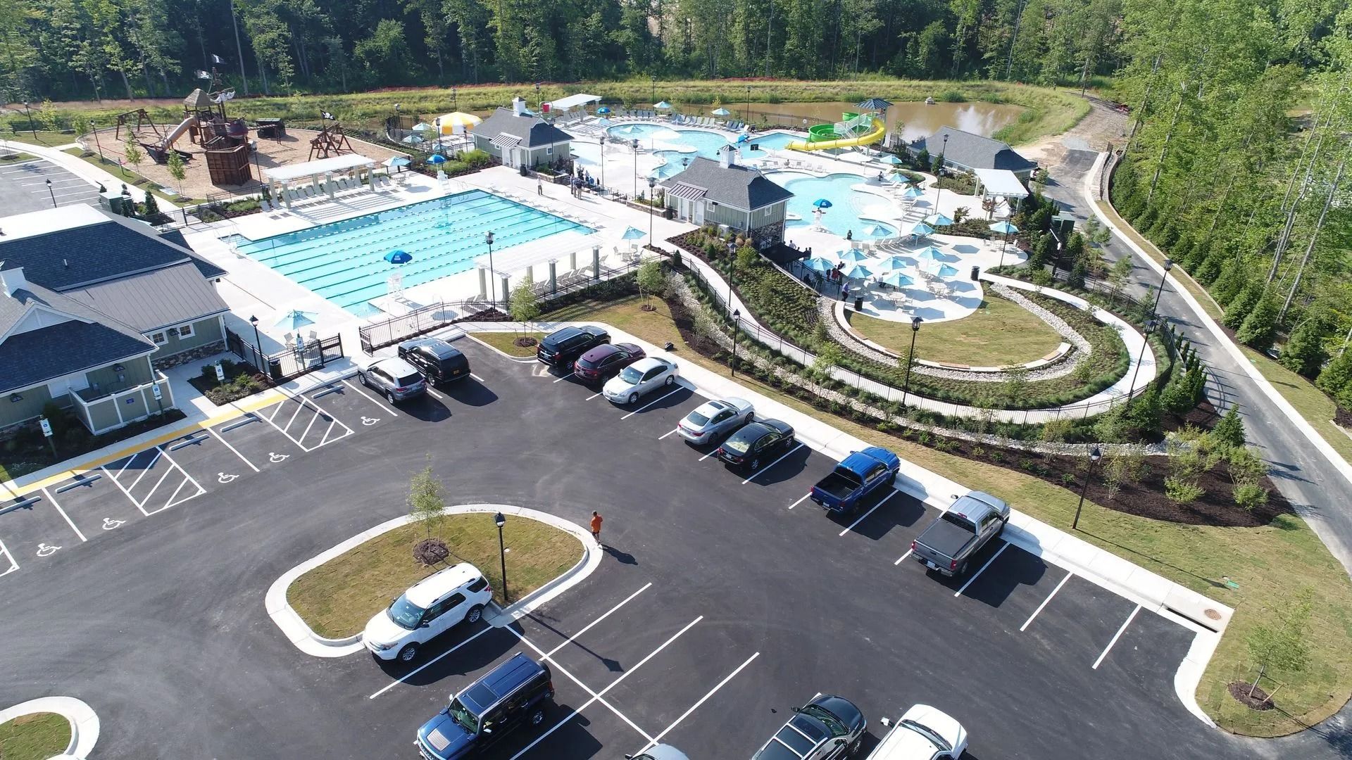 Aerial view of an outdoor pool complex with parking lot, umbrellas, and surrounding trees
