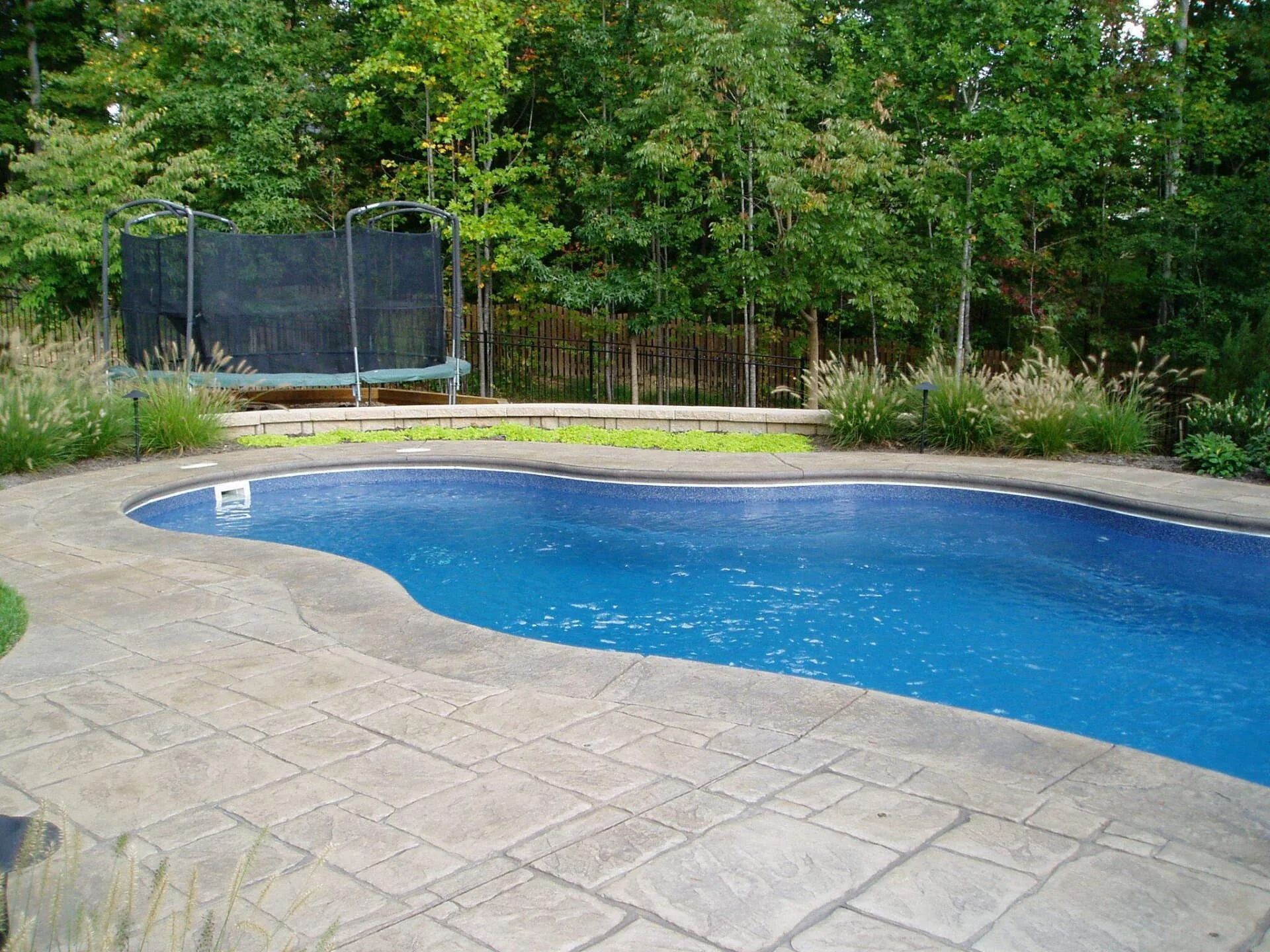 Swimming pool with stamped concrete edging, trampoline in the background, and trees.