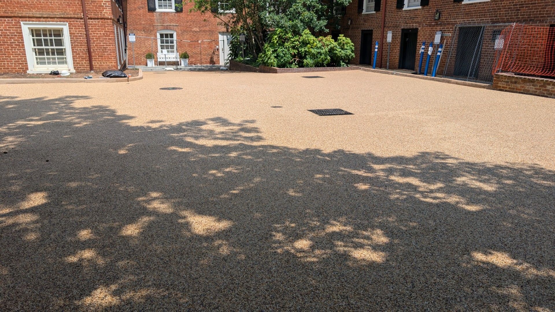 Sunlit gravel courtyard beside brick buildings, with tree shadows across the ground.