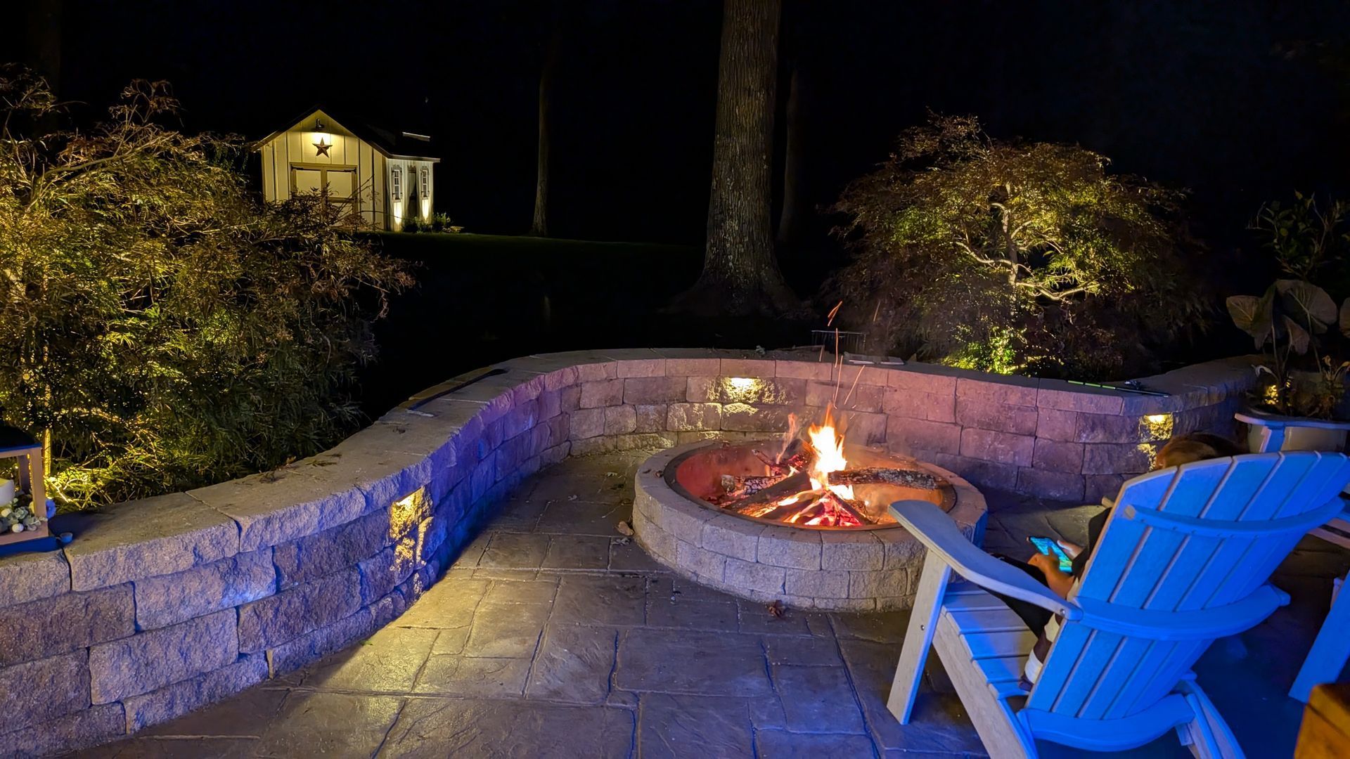 Backyard fire pit at night with blue chairs and stone seating, lit by warm flames and purple accent lights