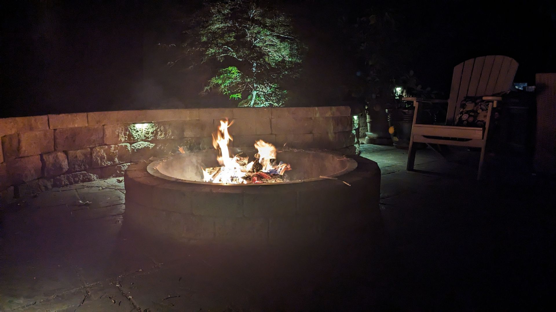 Backyard fire pit glowing at night beside a chair and low stone wall