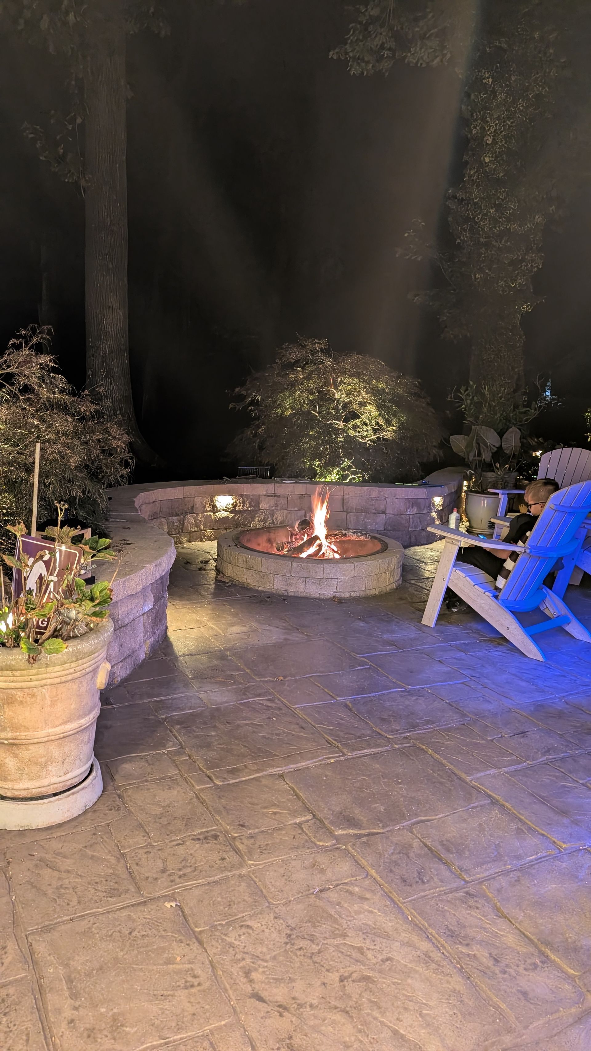 Outdoor patio at night with a stone fire pit, potted plants, and a blue lounge chair lit by warm lights