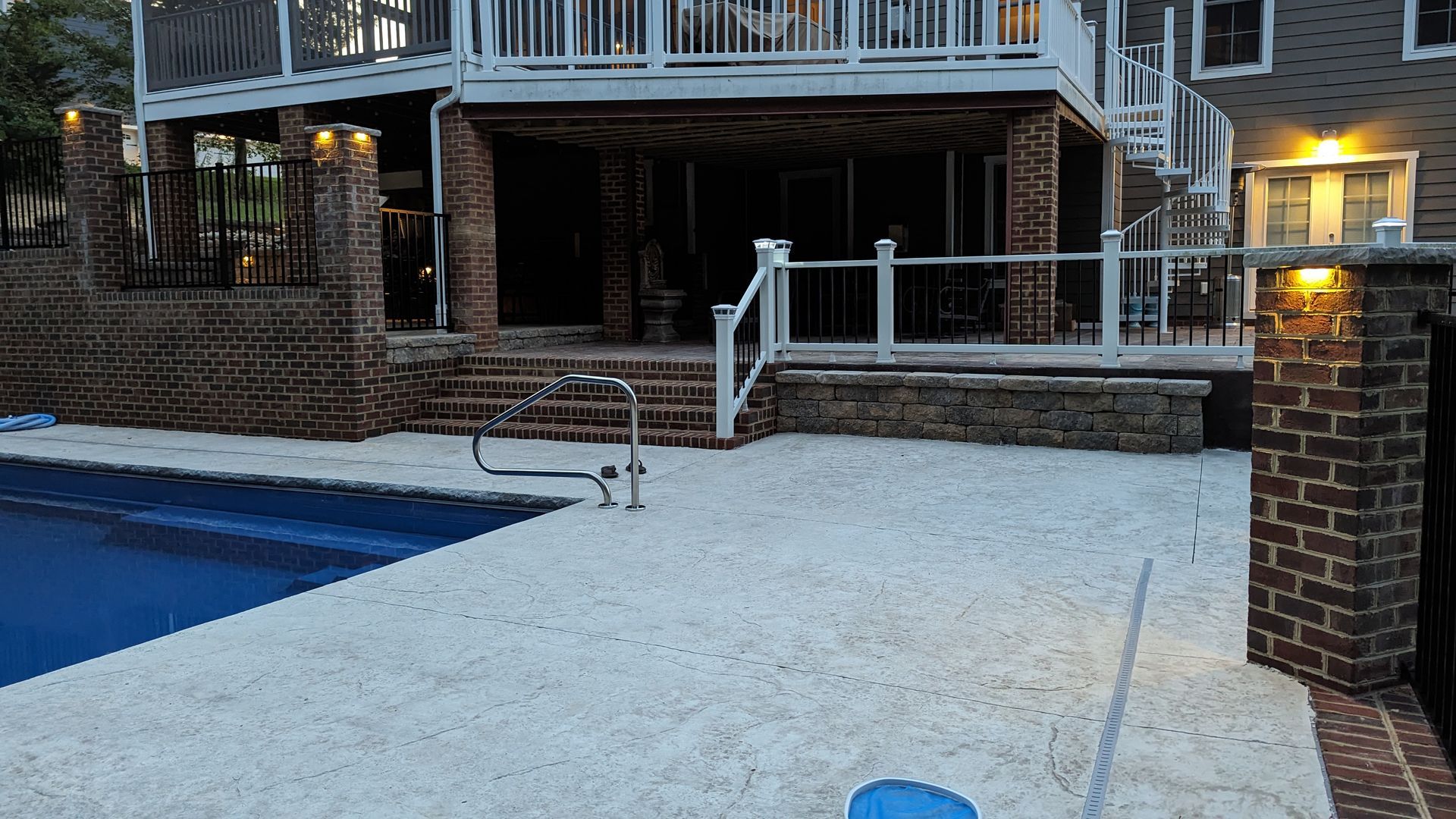 Backyard patio with white gravel, blue pool, brick pillars, and a wooden deck at dusk