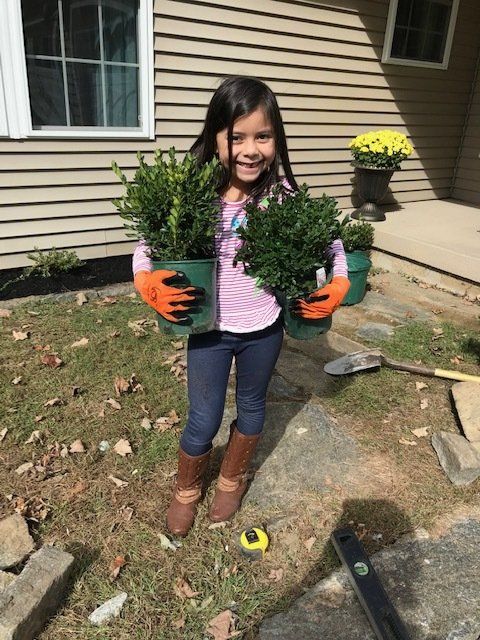 Girl smiling, holding two potted shrubs while gardening outside. Wearing gloves, boots, and jeans.