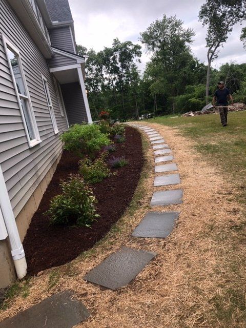 Pathway of stepping stones, mulch-lined flower bed, and a person raking grass near a house.