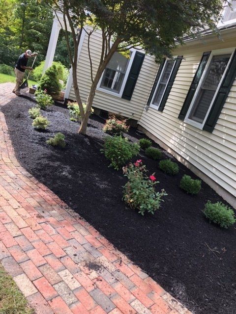 A person mulches a flower bed beside a yellow house with a brick pathway.