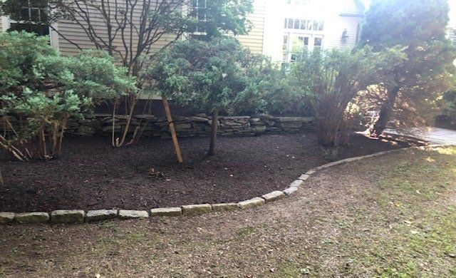 A garden bed with mulch and small trees, bordered by stones and a gravel path. House in background.
