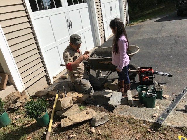 Man and young girl building with stone near garage. Man kneels, girl watches. Tools and wheelbarrow nearby.