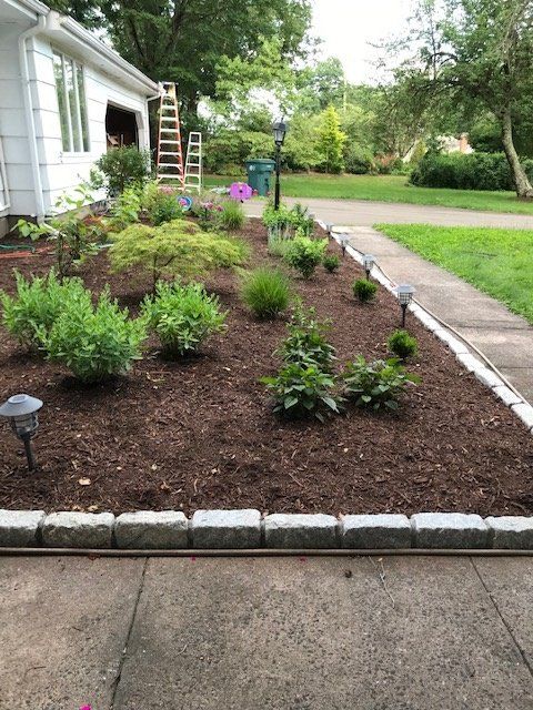 A landscaped flower bed edged with stone, next to a sidewalk. Contains green bushes, flowers, and brown mulch.