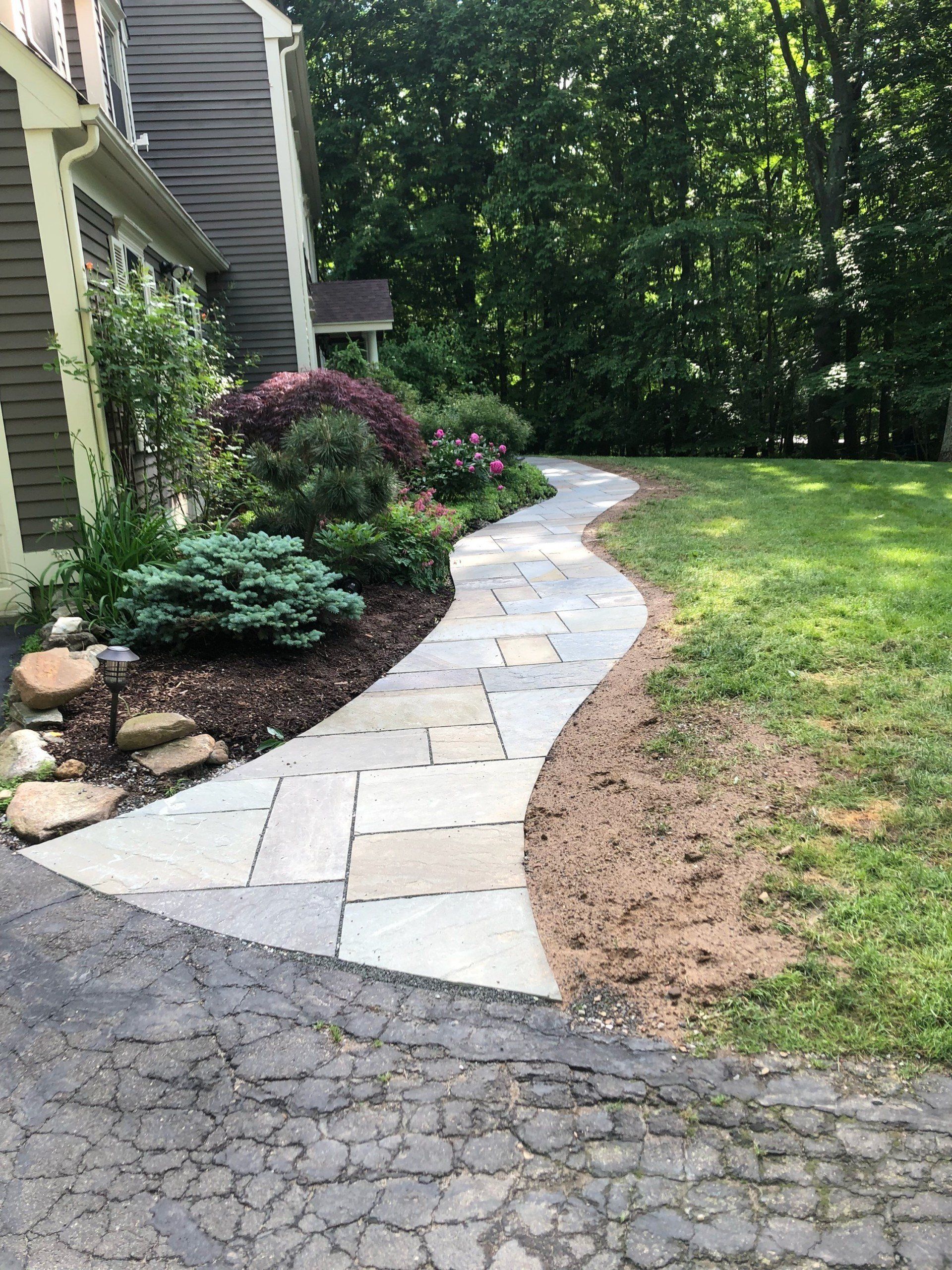 Stone walkway curves through a landscaped garden to a house. Lush green lawn to the side.