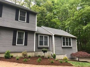 Gray house with black shutters and landscaping, surrounded by trees.