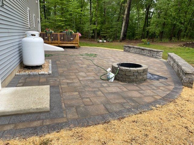Backyard patio with fire pit, propane tank, and low stone walls; gray siding and deck visible.
