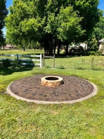Circular brick patio with fire pit, in grassy backyard with white fence and trees.