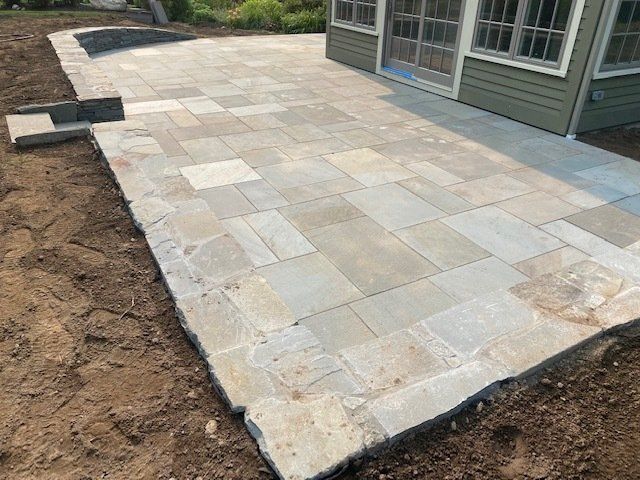 Stone patio with irregular edges and beige flagstones, next to a house with green siding.