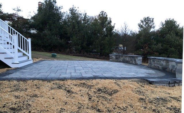 A paved patio bordered by a low wall and steps, set in a yard with dead grass and trees.