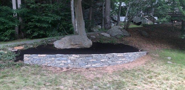 Stone retaining wall around a tree with dark mulch, set in a grassy yard with forest backdrop.
