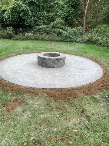 Circular stone patio with a fire pit, in a grassy backyard, trees in the background.