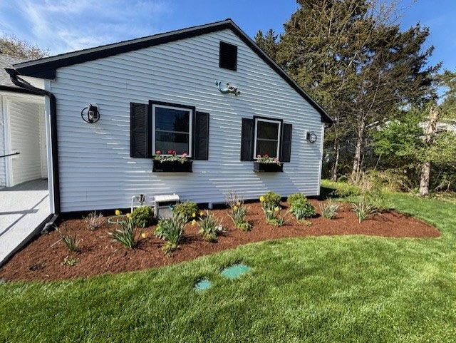 White house with black shutters and a flower bed with mulch and flowers, on a sunny day.