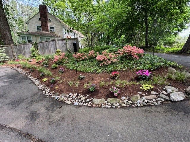 Landscaped garden bed with pink and purple flowers, mulch, and rocks in front of a house.