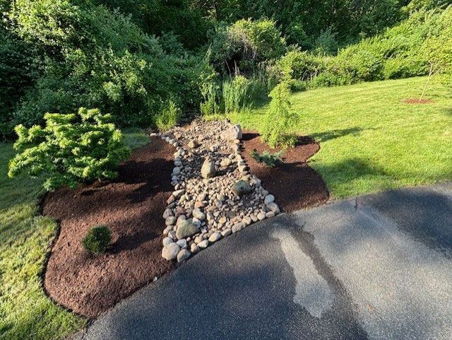 A landscaped garden bed with mulch, rocks, and small plants bordering a driveway and lawn.