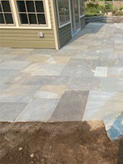 Blue-gray stone patio next to a light green house with windows and doors. Brown earth in foreground.