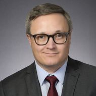 Headshot of a man in glasses, gray suit, and red tie against a gray background