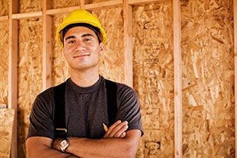 A man wearing a hard hat and suspenders is standing in front of a wooden wall.