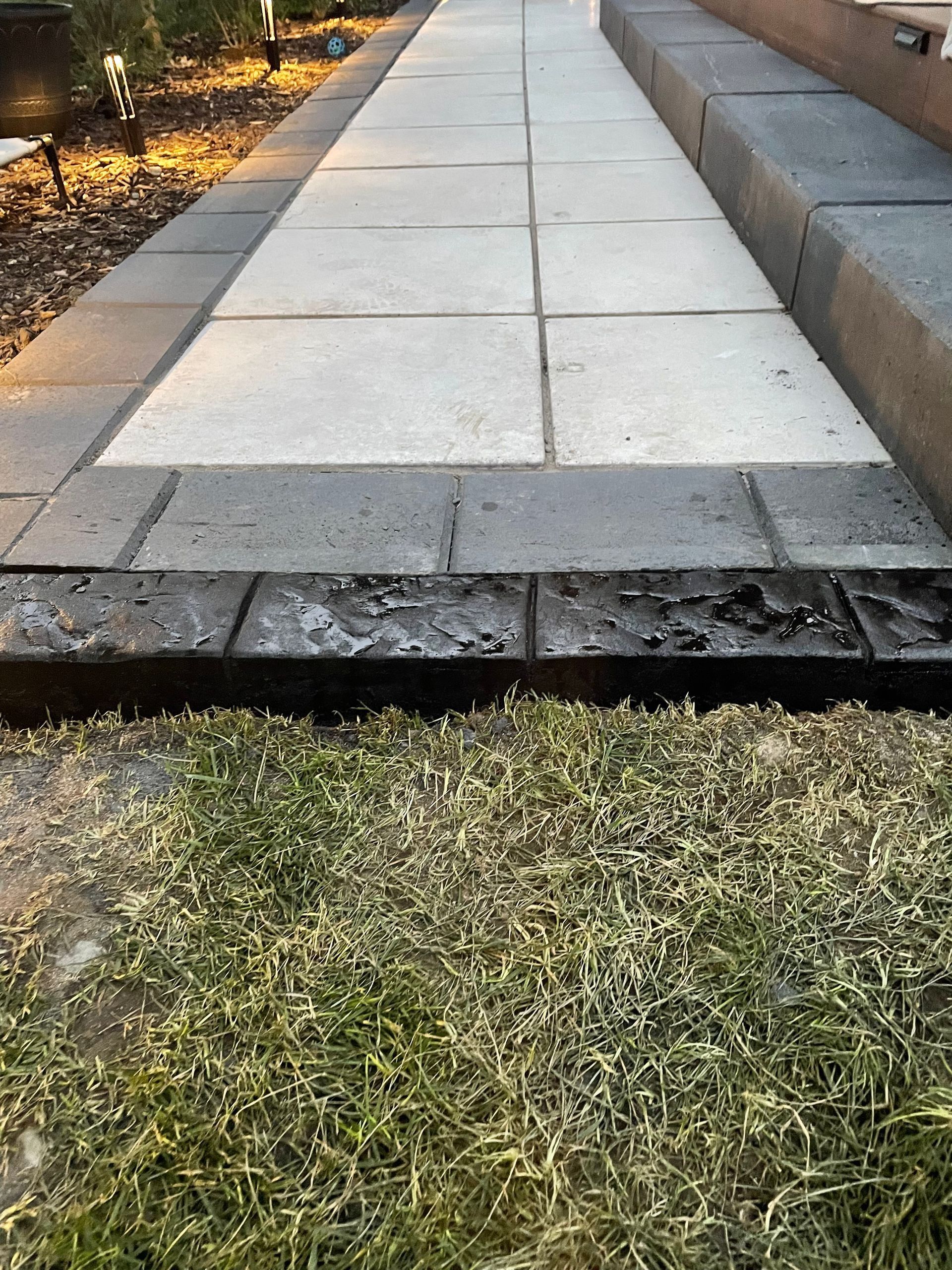Paved sidewalk with a broken edge and missing curb sections beside grass and a stone wall