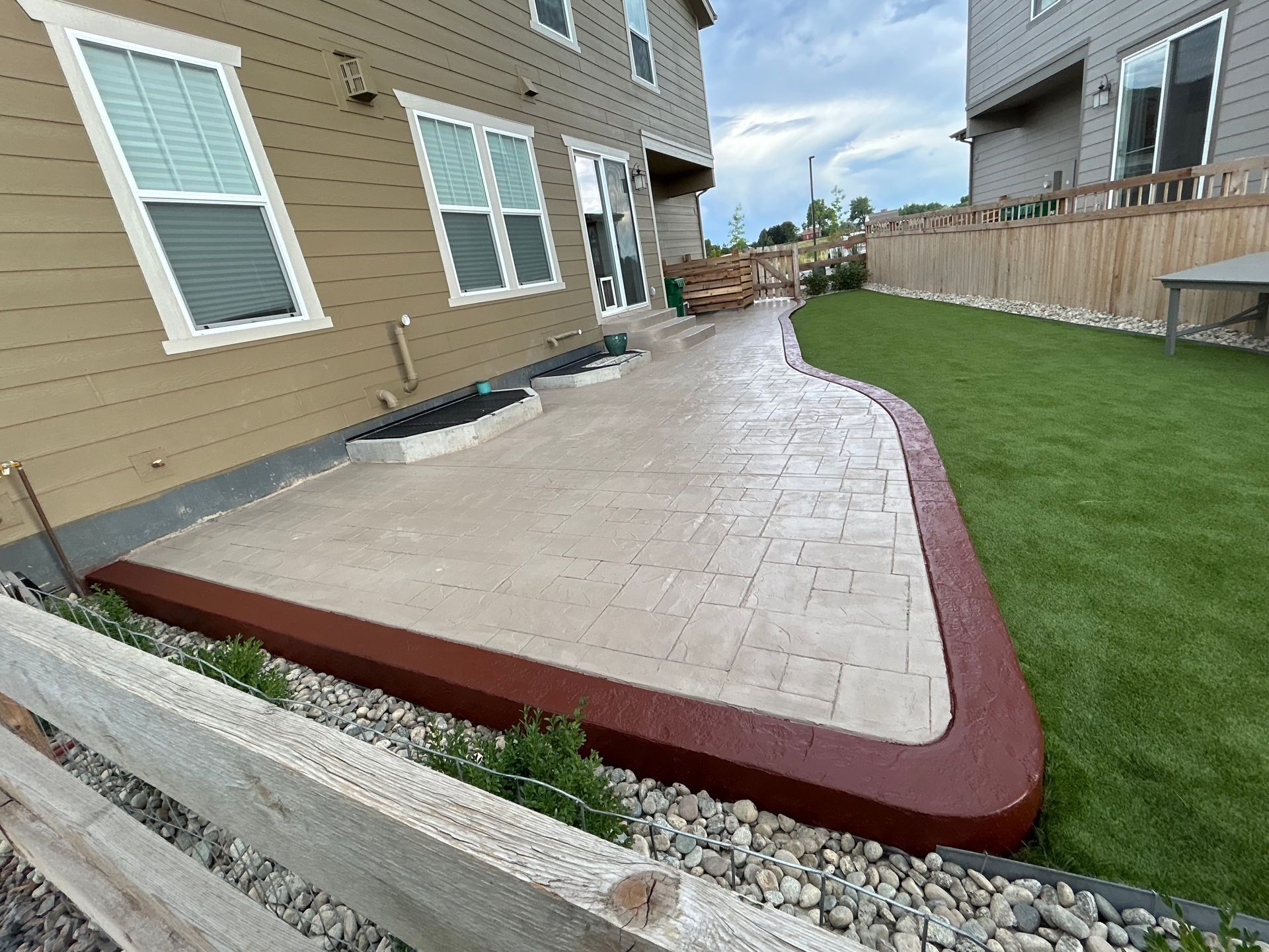Backyard concrete patio with red border beside a beige house and green lawn